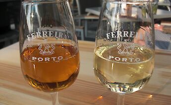 Two glasses of port wine, one amber and one light yellow, on a wooden table. The background shows blurred outdoor seating in Portugal.
