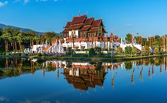 Ornate Thai architecture with red roofs reflected in a pond, surrounded by lush greenery and a clear blue sky in Chiang Mai, Thailand.