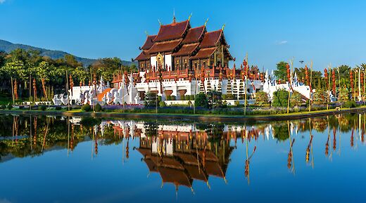 Ornate Thai architecture with red roofs reflected in a pond, surrounded by lush greenery and a clear blue sky in Chiang Mai, Thailand.