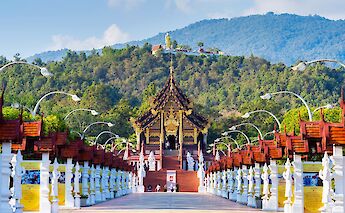 An ornate Thai pavilion with intricate designs is at the end of a grand walkway lined with trees and decorative lamp posts, with a large Buddha statue visible on a forested hillside in the background in Chiang Mai, Thailand.