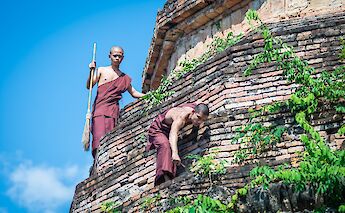 Two monks in traditional robes are engaged in cleaning tasks on an ancient brick structure in Chiang Mai, Thailand, surrounded by greenery under a blue sky.