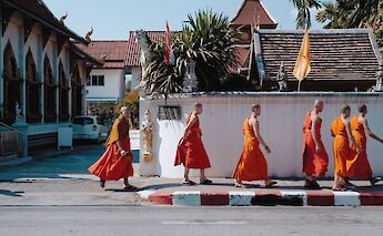 Monks in orange robes walking along a sidewalk near a temple in Chiang Mai, Thailand.