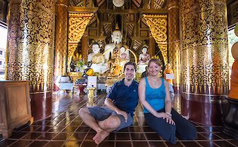 Inside a temple in Chiang Mai, Thailand, two people sit on the floor surrounded by ornate golden decorations and Buddha statues.