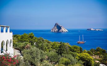 A scenic coastal view from Italy, featuring a white building with arches, lush greenery, a clear blue sea, and a distinct rocky outcrop in the distance, with a sailboat on the water.