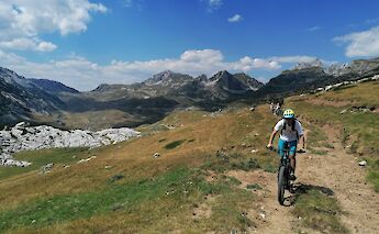 A cyclist wearing a helmet rides on a grassy trail with rugged peaks in the background, in Zabljak, Montenegro.