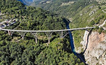 Aerial view of a large bridge spanning a deep green valley with a river below, surrounded by forested mountains.
