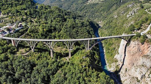 Aerial view of a large bridge spanning a deep green valley with a river below, surrounded by forested mountains.
