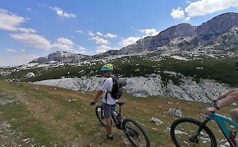 Cyclists pause on a trail overlooking rugged mountainous terrain with rocky outcrops under a bright blue sky in the Durmitor region of Montenegro.