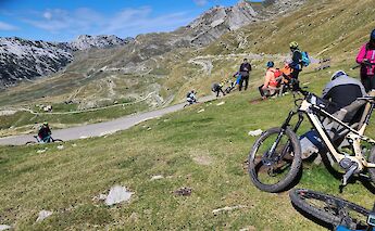 Cyclists resting on a grassy hill with winding roads and mountain views in the background.