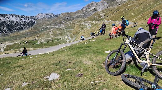Cyclists resting on a grassy hill with winding roads and mountain views in the background.