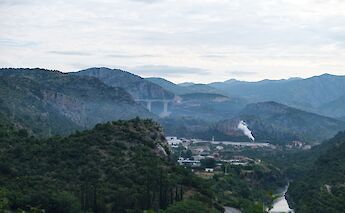 View of a valley with distant mountains, a large bridge, and a small town emitting smoke.