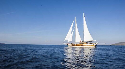 A traditional sailing yacht with full white sails, cruising on open sea under a clear blue sky. Hills or islands are visible in the distant background.