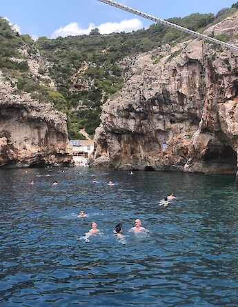 People swim in a secluded cove with rocky cliffs and lush greenery in Northern Dalmatia, Croatia. The clear blue water contrasts with the rugged landscape.