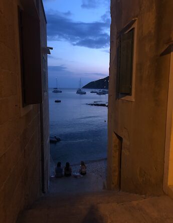 A serene view through narrow stone alleyways opens to a nighttime seascape with anchored boats in Northern Dalmatia, Croatia. Three people sit at the water's edge, enjoying the evening atmosphere.