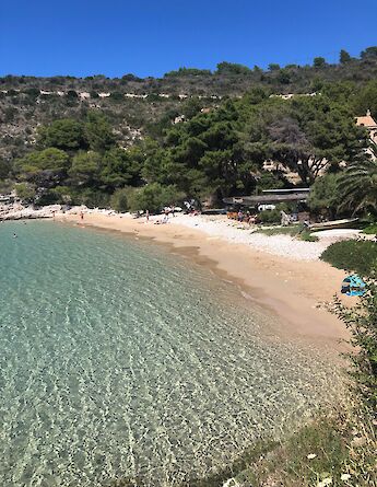 A quiet, sandy beach with clear, shallow waters and gentle waves is nestled against a tree-covered hillside in Northern Dalmatia, Croatia. Visitors enjoy the tranquil setting.