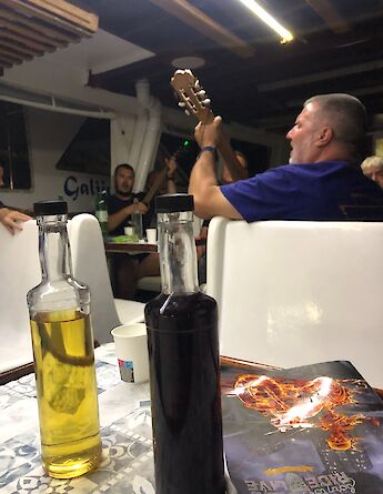 Indoor setting with people playing guitars in a casual gathering; bottles of liquid are on a table in the foreground.