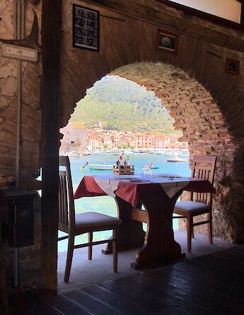 A cozy dining setup for two under a rustic stone archway, offering a view of a seaside town in Croatia.