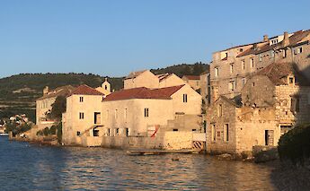 Stone buildings with red-tiled roofs line the waterfront in a coastal town, illuminated by warm evening sunlight. Hills covered with greenery rise in the background, and the calm waters reflect the golden hues of the setting sun. This scene is from northern Dalmatia, Croatia.