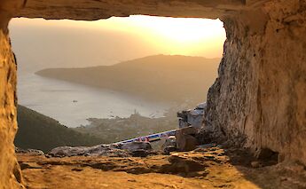 A scenic view framed by a stone opening shows a coastal landscape at sunset in Northern Dalmatia, Croatia. The soft, golden light illuminates the distant shoreline and hills.