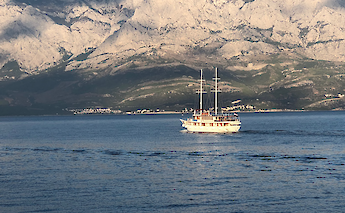 A sailboat with masts sails on a blue sea with a backdrop of rugged, mountainous terrain in Croatia.