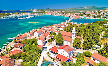 Aerial view of a coastal town in northern Dalmatia, Croatia, featuring red-roofed buildings, a marina with boats, and turquoise waters.