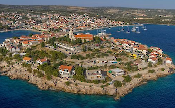 Aerial view of a peninsula in northern Dalmatia, Croatia, with a cluster of red-roofed buildings, a church with a bell tower, and surrounding blue waters.