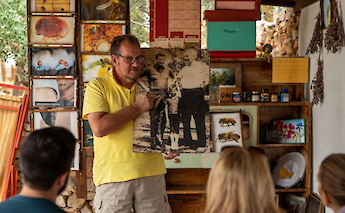 A man in a yellow shirt holds a historical photograph, presenting it to a small group. Behind him, shelves display jars, beekeeping imagery, and dried herbs, suggesting a local cultural or beekeeping presentation in Croatia.