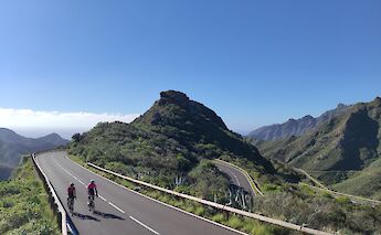 Two cyclists on a curving mountain road surrounded by lush green hills under a clear sky in Tenerife, Spain.