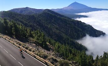 A cyclist on a mountain road with lush green hills and Mount Teide visible in the distance in Tenerife, Spain.