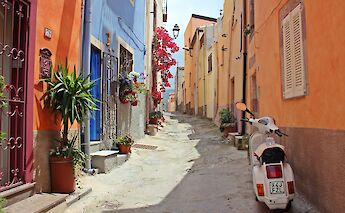 A narrow alley in Sardinia, Italy, flanked by colorful buildings with potted plants and a parked scooter.
