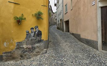 Cobblestone street in Oristano, Sardinia, Italy, with a mural of three people on a yellow wall and small potted plants.