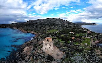 An elevated view of a coastal landscape in Tharros, Sardinia, showcasing a rugged shoreline, blue waters, and a historical stone tower.
