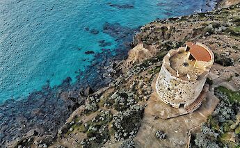 An aerial view of a historic stone tower on a cliff beside the turquoise sea, surrounded by rocky vegetation in Sardinia, Italy.