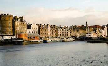 A view of boats docked along the waterfront in Leith, Edinburgh, Scotland, with historic stone buildings in the background.
