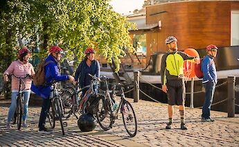 A group of cyclists with bicycles and helmets standing on a cobbled path by the Union Canal in Edinburgh, Scotland, with a wooden building and trees in the background.