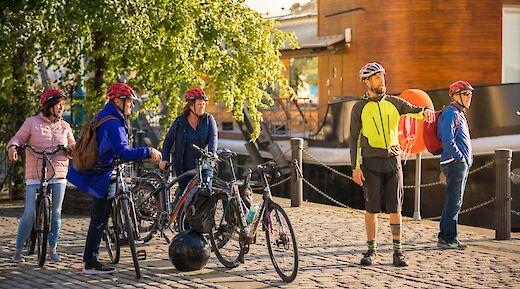 A group of cyclists with bicycles and helmets standing on a cobbled path by the Union Canal in Edinburgh, Scotland, with a wooden building and trees in the background.
