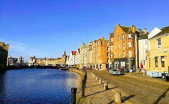 A canal view in the Shore of Leith, Edinburgh, Scotland, with historic stone buildings lining the waterside under a blue sky.