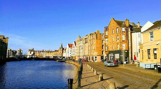 A canal view in the Shore of Leith, Edinburgh, Scotland, with historic stone buildings lining the waterside under a blue sky.