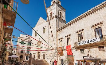 A sunny square in Putignano with historic stone buildings, colorful banners, and seating areas.
