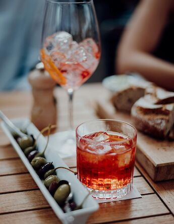 Two glasses of cocktails, a dish of assorted olives, and a plate of bread on a wooden table.