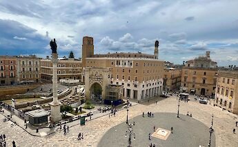 Piazza Sant'Oronzo with a Roman amphitheater and surrounding buildings in Lecce, under a cloudy sky.