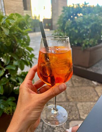 A hand holding a glass of Aperol Spritz outdoors, with plants and a stone pavement in the background.