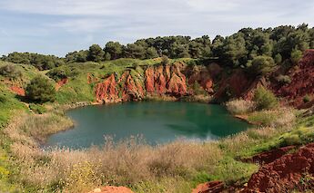 A vivid red and green landscape with a small lake surrounded by red bauxite cliffs and greenery at the Cave di Bauxite near Otranto, Italy.