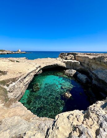 A rocky coastal area with clear turquoise water and a natural arch, set against a backdrop of the deep blue sea in Salento, Italy.