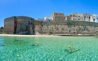 A stone fortress wall along a clear, turquoise shoreline in Salento, Italy, with buildings atop the wall under a clear blue sky.