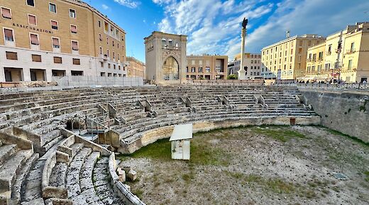 The Roman Amphitheater in Lecce, Italy, surrounded by buildings under a partly cloudy sky.