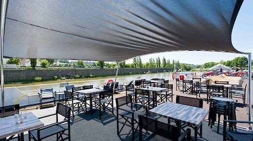 A shaded outdoor dining area on the deck of a boat with tables and chairs overlooking a river and distant greenery.