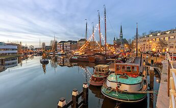 A harbor scene in Gouda, Holland, with moored boats, historic buildings, and twinkling lights reflected on the water.