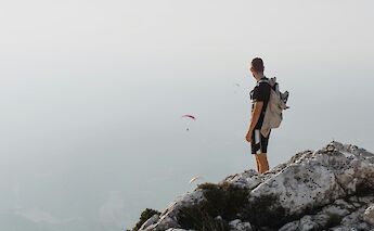 A person standing on a rocky outcrop, gazing at paragliders in the hazy sky, Montagne Sainte-Victoire area, Aix-en-Provence, France.