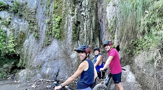Three people with mountain bikes are posing in front of a waterfall, surrounded by lush greenery and rocky terrain.